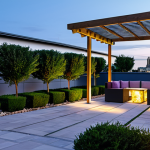 A beautifully designed, modern rooftop garden in a European city, featuring lush Mediterranean plants like lavender and rosemary, interspersed with elegant climbing roses. Natural stone pathways wind through the space, leading to a sophisticated seating area with a contemporary pergola. A subtle, serene water feature provides a calming ambiance, and discreet solar lights illuminate the area as dusk approaches. The urban skyline is visible in the background. The image should convey professional architectural photography, with perfect anatomy, correct proportions, and a natural composition. safe for work, appropriate content, fully clothed, professional.